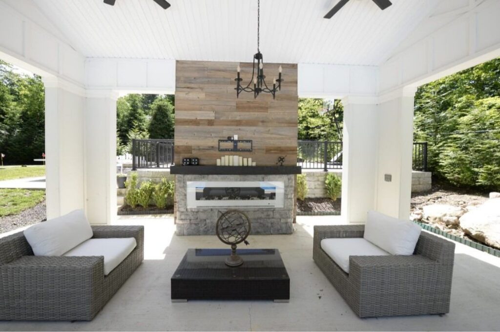 13-2048x1361 Outdoor patio with wicker sofas, stone fireplace, wooden wall, and hanging chandelier.