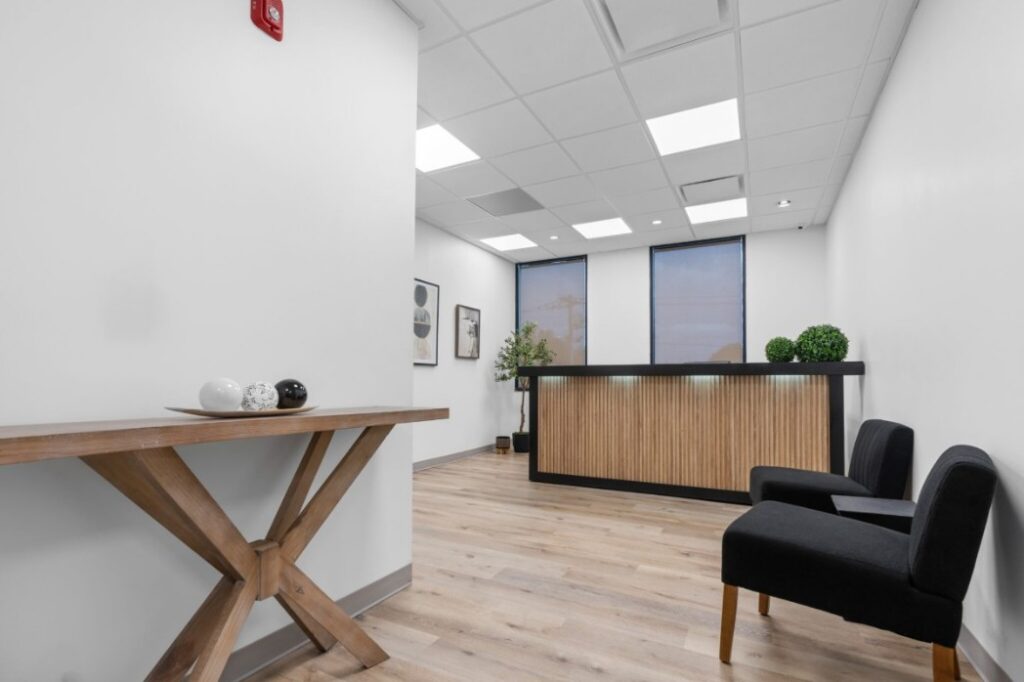 Ikon-2-10-2048x1365 Modern reception area with wooden desk, black chairs, abstract art, and plants on hardwood floor.