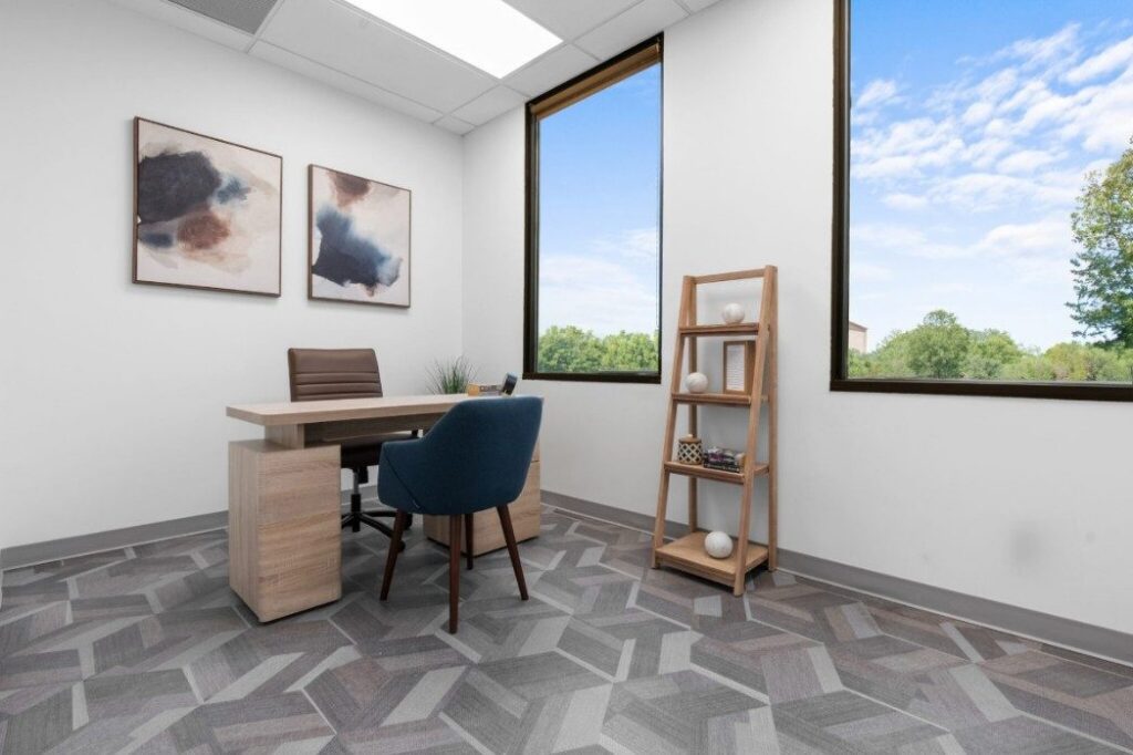 Ikon-2-4-2048x1365 Modern office with abstract paintings, desk, blue chair, shelf, and large window showing trees outside.