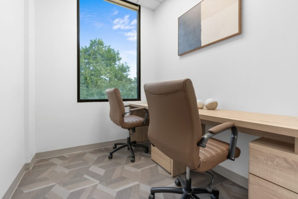 Ikon-6-2048x1365 Two brown office chairs in a minimalistic room with a wooden desk and large window view of trees.
